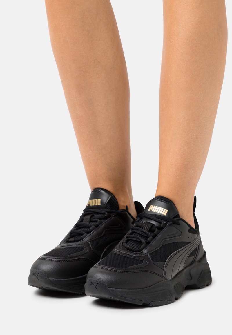Paire de baskets noires Puma portées aux pieds avec les parties inférieures des jambes visibles, montrant une semelle épaisse et un logo Puma doré sur la languette.