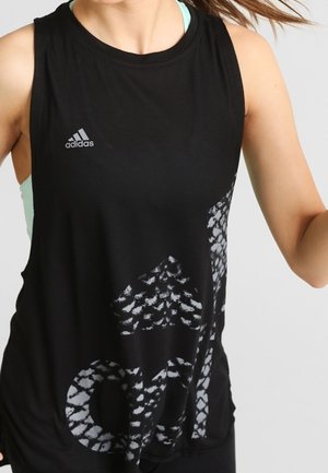 Kvinde iført en sort Adidas ærmeløs top med stort mønstret Adidas-logo og lyseblå sports-bh underneath.