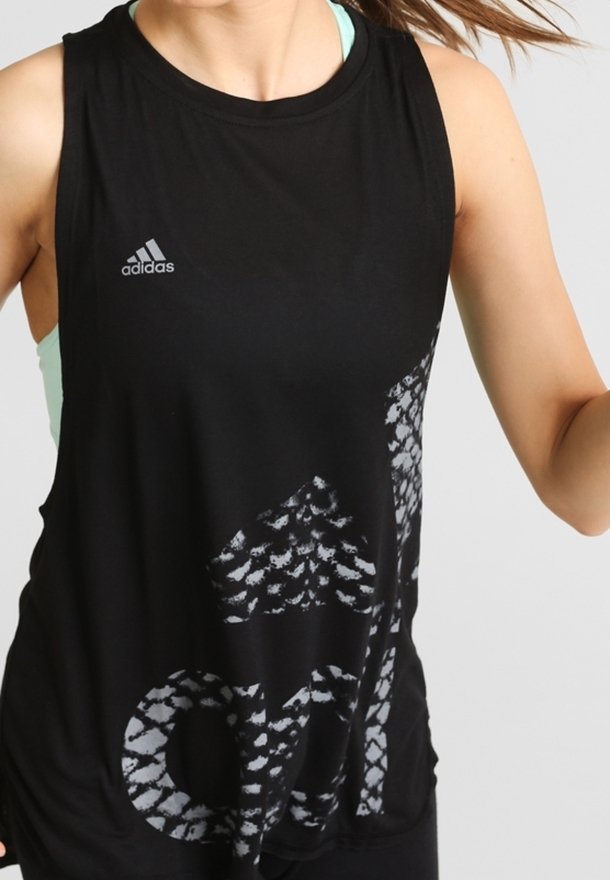 Femme portant un débardeur noir Adidas avec un grand logo Adidas à motif et un soutien-gorge de sport bleu clair en dessous.