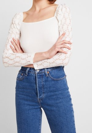 Witte top met lange mouwen en kanten pofmouwen, gecombineerd met een high-waisted blauwe spijkerbroek. De top heeft een vierkante halslijn en een aansluitend lijfje.