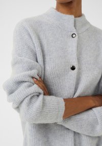 Hellblauer, gerippter Cardigan mit hohem Kragen, ausgestattet mit einem einzelnen schwarzen Knopf und weicher, flauschiger Textur. Die Ärmel sind an den Handgelenken leicht gerafft.