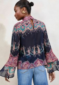 Zračna temno modska bluza s paisley vzorcem v turkizni, roza in zlati akcentih. Ima zvonaste rokave in zadaj gumbasto zapiranje.