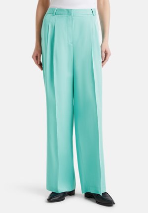 Pantalones de pierna ancha en color turquesa, confeccionados en una tela ligera, con pliegues frontales y pasadores para cinturón, combinados con zapatos planos negros.