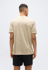 Άνδρας που φαίνεται από πίσω φοράει μπεζ κοντομάνικο μπλουζάκι Under Armour και μαύρο σορτς, με φόντο έναν απλό ανοιχτόχρωμο τοίχο.