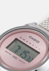 Digital klocka från Casio med rund silverfärgade boett, rosa urtavla och nätarmband. Visar tiden med vattentålighet angiven längst ner.