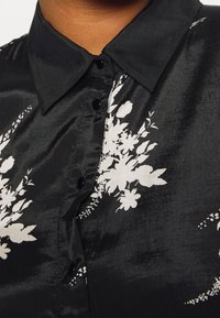 Camicia di raso nera con colletto, decorata con motivi floreali bianchi, bottoni neri e una texture liscia.