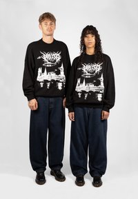 Svarte strikkede hettegensere har et hvitt grafisk design av en bysilhuett og UFOer. Begge modellene har på seg løstsittende denimbukser og svarte sko.