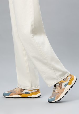 Personne portant un pantalon blanc à jambes larges et des baskets multicolores beige, bleu et jaune, marchant sur un fond gris.