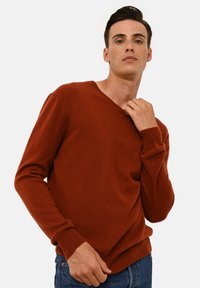 Maglione a V colore ruggine realizzato in un morbido tessuto a maglia, con maniche lunghe e polsini a coste, abbinato a jeans in denim blu.
