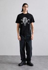 Svart bomull t-skjorte med et sølvfarget grafisk trykk av et kranium og en iskremkjegle. Kombinert med løse mørke jeans og svarte sko.