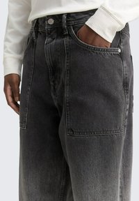 Osoba o ciemnej karnacji, ubrana w czarne dżinsy i białą koszulę z długim rękawem, z ręką częściowo w kieszeni dżinsów.