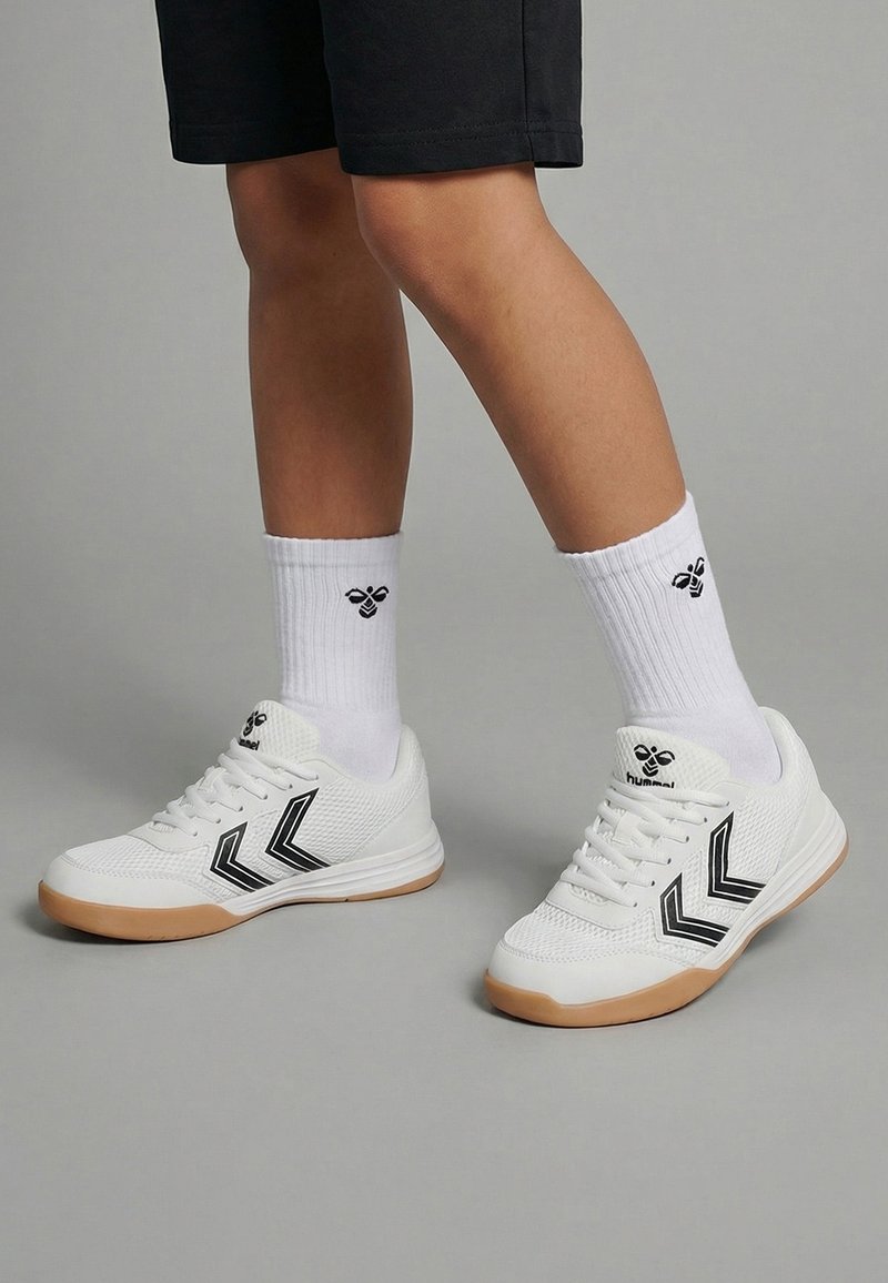 Benedenbenen met witte sportschoenen met zwart chevronpatroon en witte sokken met zwart bijenlogo, op een grijze vloerachtergrond.