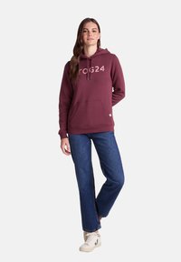 Maroon hooded sweatshirt with front kangaroo pocket, graphic logo "TOG24" in light color, paired with blue wide-leg jeans and light sneakers.