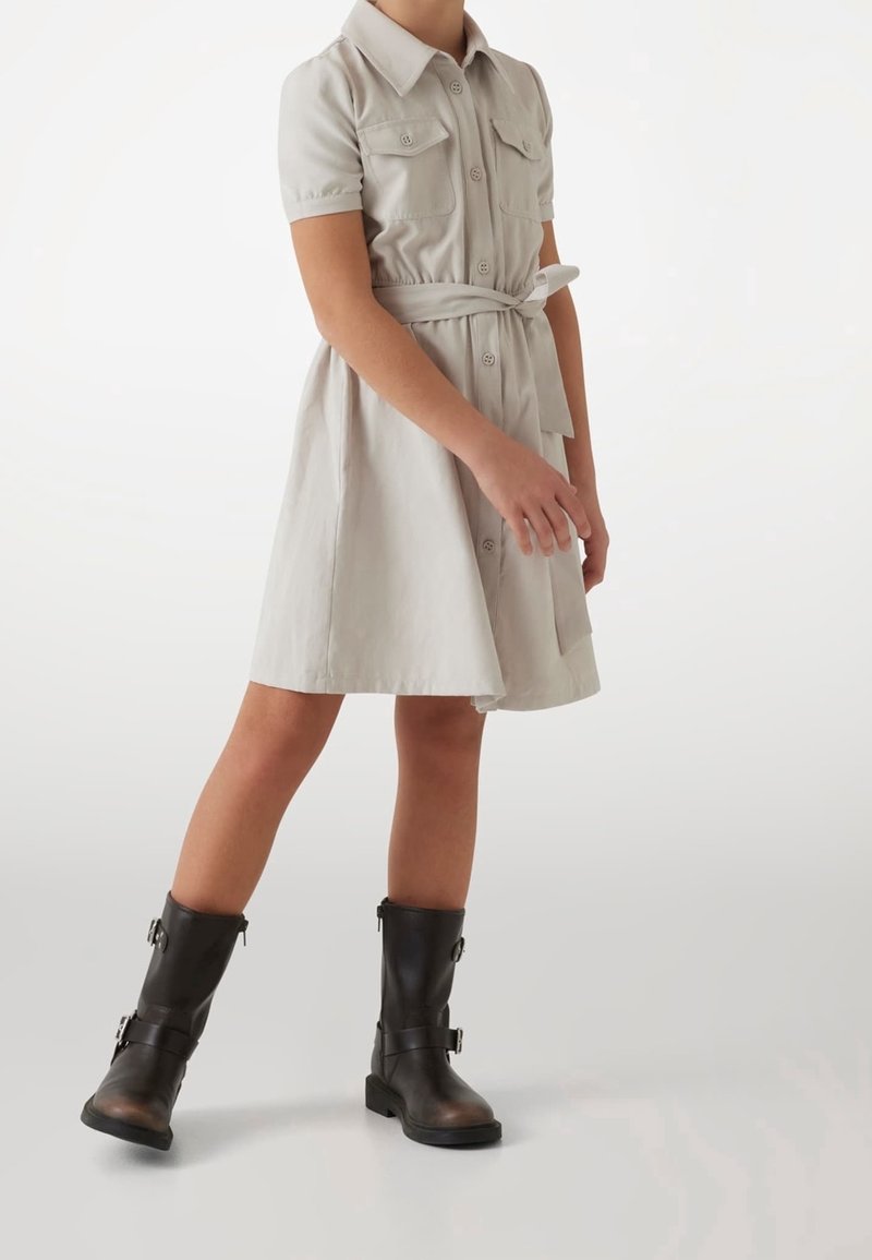 Jasnoszara sukienka koszulowa z krótkimi rękawami, zapięciem na guziki, dwoma kieszeniami na piersi i paskiem w talii. Połączona z ciemnobrązowymi kozakami sięgającymi kolan.
