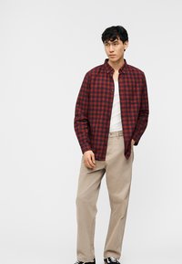 Νεαρός άντρας που στέκεται φορώντας κόκκινο και μαύρο πουκάμισο με κουμπιά πάνω από λευκό μπλουζάκι, μπεζ παντελόνι και μαύρα αθλητικά παπούτσια, σε απλό φόντο.