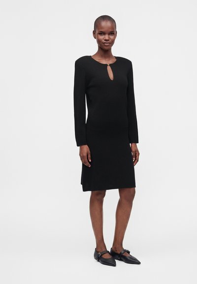 Femme portant une robe noire à manches longues jusqu'au genou avec un petit décolleté en forme de serrure et des ballerines noires pointues avec des brides.