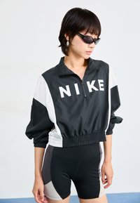 Jachetă Nike alb-negru, din material ușor, cu guler zip și tiv elastic, purtată peste pantaloni scurți sport negri și gri. Design elegant și modern.