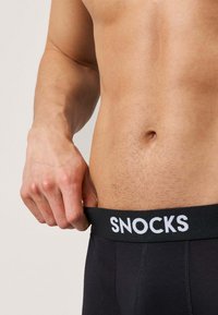 Biancheria intima da uomo nera con una fascia larga in vita con la scritta bianca "SNOCKS". Il tessuto sembra liscio ed elastico.