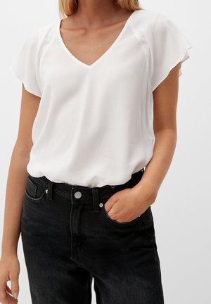 Vrouw draagt een witte blouse met korte mouwen en V-hals, ingestopt in een zwarte high-waisted jeans, met één hand in het voorzakje.