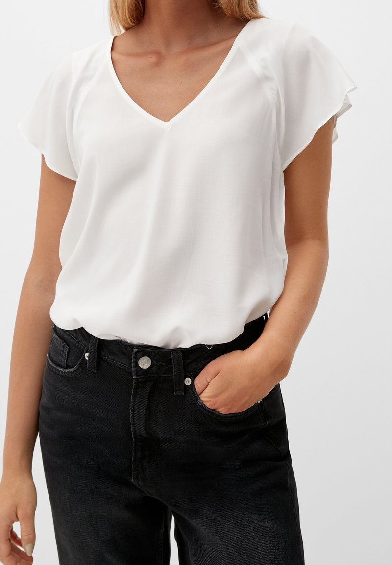 Femme portant un chemisier blanc à manches courtes et col en V, rentré dans un jean noir taille haute, une main dans la poche avant.