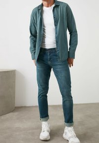 Camisa de mezclilla en verde azulado desgastado, abotonada con cuello; combinada con jeans azules oscuros, con puños arremangados, y zapatillas blancas. Tejido suave, corte casual.