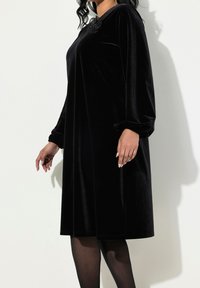Robe en velours noir à manches longues, avec une coupe décontractée et des poignets froncés. Présente un accent floral au niveau de l'encolure. Texture lisse.