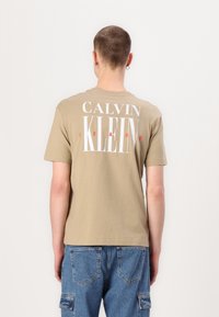 Beige bavlněné tričko s krátkým rukávem a kulatým výstřihem, na zadní straně s velkým bílým a červeným nápisem "CALVIN KLEIN JEANS."