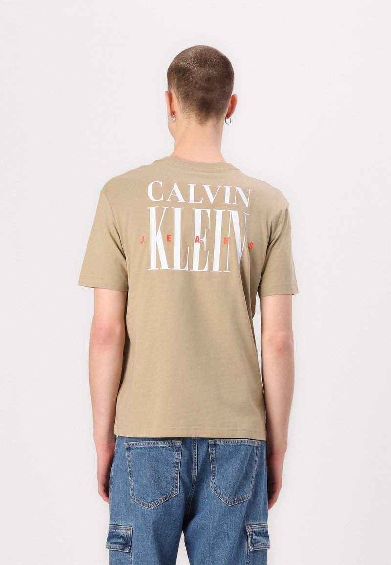 Beige bavlněné tričko s krátkým rukávem a kulatým výstřihem, na zadní straně s velkým bílým a červeným nápisem "CALVIN KLEIN JEANS."