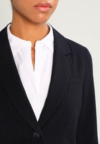 Marinblått pinstripe-blazer med skräddarsydd passform, enkel knappstängning och skarpa slag, lager-på-lager över en vit skjorta med krage.