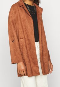 Brun faux suede frakke med notched krave, åben front, sidelommer og justerbare ærmer. Sammen med en sort top og lyse bukser.