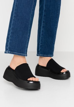 Fødder iført sorte platform-slide sandaler med åbne tæer og mørkeblå cropped jeans mod en ensartet baggrund.