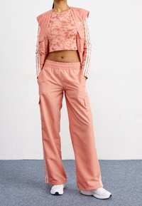 Person som bär en laxrosa Adidas träningsjacka, en tie-dye crop top, vida byxor och vita sneakers medan de står på en blå matta.