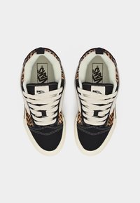 Ζευγάρι μαύρα και λευκά παπούτσια Vans με λεπτομέρειες σε λεοπάρ μοτίβο και λευκά κορδόνια, εμφανιζόμενα από πάνω σε απλό φόντο.