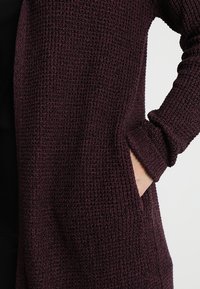 Cardigan en maille bordeaux présentant un motif texturé, des manches longues et des poches latérales. Le tissu semble doux et confortable.