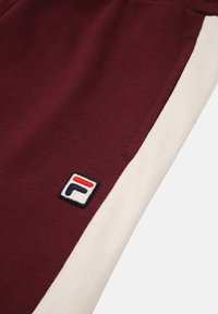 Bordeauxkleurige sweatpants met een crèmekleurige zijkant, gemaakt van een gestructureerde stof en voorzien van een geborduurd logo in rood, wit en blauw op de bovenbeen.