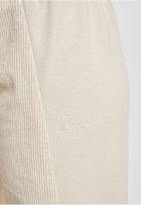 Beige Baumwollmischungs-Hosen mit gerippter Textur an der Seite und einem weißen gestickten Logo. Das Material wirkt weich und lässig.
