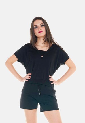 Jonge vrouw met lang bruin haar, gekleed in een losse zwarte top en korte broek, staat met haar handen in haar zij tegen een witte achtergrond.