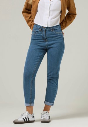 Kobieta w krótkich niebieskich dżinsach, białej koszuli zapinanej na guziki, brązowej kurtce i białych trampkach z czarnymi paskami, stojąca z rękami za plecami.