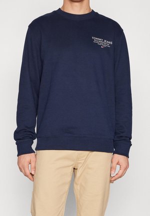 Homme portant un sweat-shirt Tommy Jeans bleu marine avec un texte blanc sur la poitrine et un pantalon beige, debout devant un fond uni.