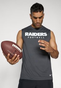 Siva brezrokavnik s tekstom "RAIDERS FOOTBALL", ki drži rjavo nogometno žogo, na enobarvni svetli podlagi. Na srajci je logotip Nike.