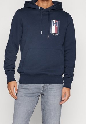 Mężczyzna noszący granatową bluzę z kapturem z kieszenią z przodu i logo Tommy Hilfiger w kolorach czerwonym, białym i niebieskim na lewej piersi, zestawioną z jasnoszarymi jeansami.