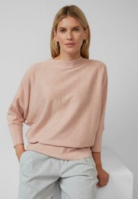 Różowy sweter o fakturze z wysokim kołnierzem i opuszczonymi ramionami, w połączeniu z jasnoszarymi spodniami. Miękki, casualowy materiał z detalami ściągaczy.