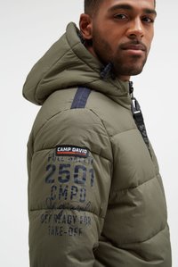 Uomo che indossa una giacca imbottita verde oliva con cappuccio, caratterizzata dalla toppa "Camp David" e da testo stampato sulla manica.