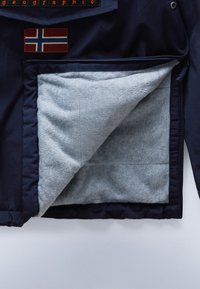Marineblaue Jacke mit grauer Fleece-Innenseite, teilweise geöffnet, zeigt roten und blauen Flaggenaufnäher und orangefarbenen bestickten Text auf der Brust.
