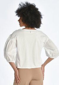 Vit kortärmad topp med puffärmar, utrustad med elastiska muddar och en enkel, ren design. Bakre nackdel inkluderar en liten etikettdetalj.