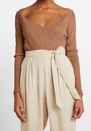 Kobieta w brązowej teksturowanej bluzce z odkrytymi ramionami, z długimi rękawami oraz beżowych spodniach z wysokim stanem, zawiązanych po bokach na kokardę.