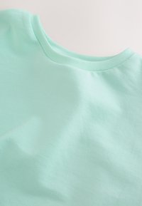 Tissu de t-shirt en coton turquoise clair à col rond, texture lisse et légères plis naturels sur un fond neutre.