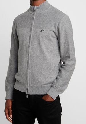 Homme portant un pull zippé gris clair avec col, poignets et bas côtelés, avec un logo A/X sur la poitrine, associé à un pantalon noir.