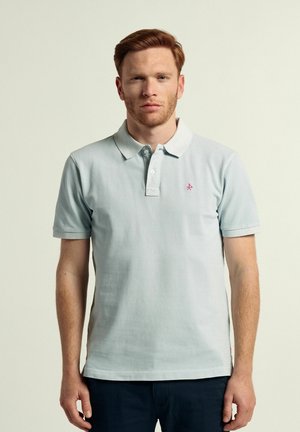 Camisa polo de color azul claro con mangas cortas, un cuello, tres botones y un pequeño logo rosa en el lado izquierdo del pecho. Textura de algodón suave.