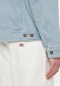 Ljusblå denimjacka med bronsknappar och en sydd fåll. Under jackan, vita byxor med en synlig Dickies-logotyp och fickor.